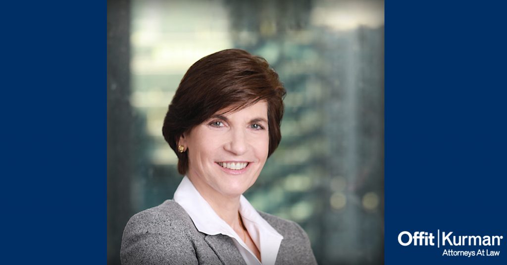 Professional headshot of Bettina Hindin, between 2 blue pillars with the Offit kurman Logo in the bottom left side of the picture