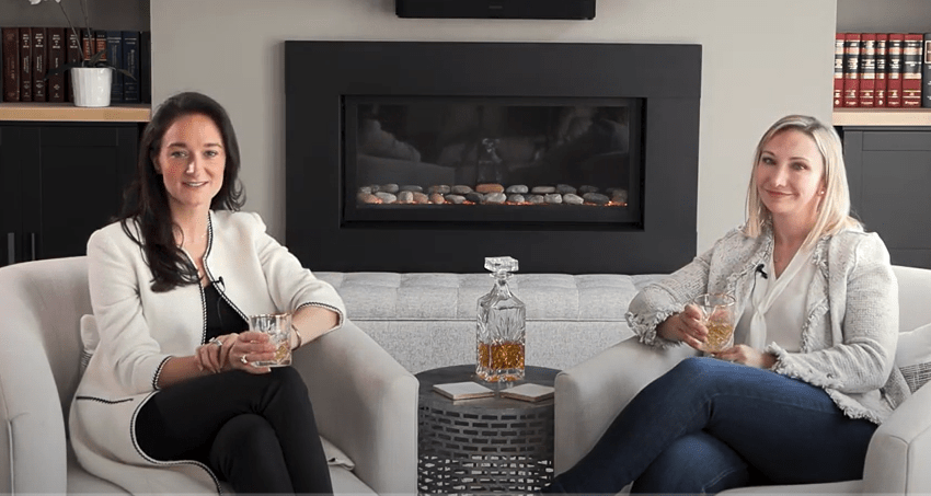 Image of Rachel Mech & Emily Shank sitting in front of an electric fireplace with drinks in their hands