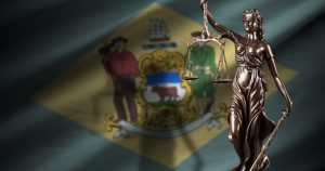 A bronze statue of Lady Justice holding scales, set against the background of the Delaware state flag
