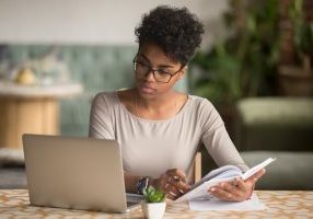 Focused young african american businesswoman or student looking at laptop holding book learning