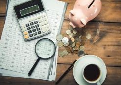 calculator and magnifying glass on top document with piggy bank, coins and a coffee cup on a wooden table