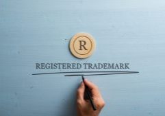 Letter R cut into wooden cut circle and male hand writing a Registered trademark sign under it. Over pastel blue wooden background.