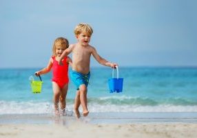 Little boy and a girl, siblings run barefoot on sea beach from waves carry toy buckets with water
