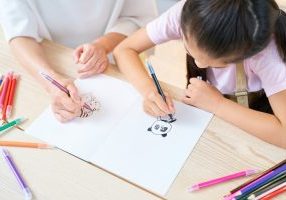 Parent and child drawing on a white piece of paper with colored pencils scattered on a wooden table