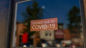 Close-up on a closed sign in the window of a shop displaying the message Closed due to Covid-19. The glass reflects the day city. 3d rendering