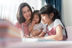 Homeschool Asian young little girls learning, reading and do homework while kind mother help and teach her. Mom and baby sister smile and encourage to girl. Girl happy to study at home education.