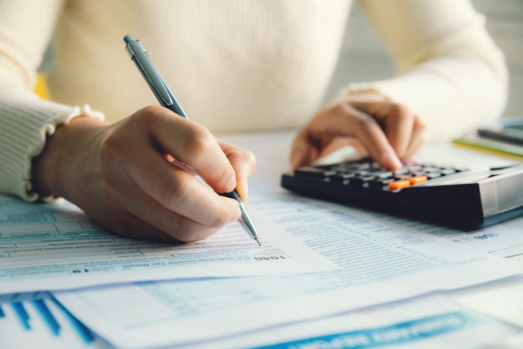 Closeup of a woman's hands filling out a 1040 tax form. She has a pencil in her right and and a calculator in her left hand.