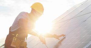 A worker wearing a hard hat and safety goggles is installing or maintaining solar panels under bright sunlight. The worker is using tools from a tool belt and is positioned in front of an array of solar panels.