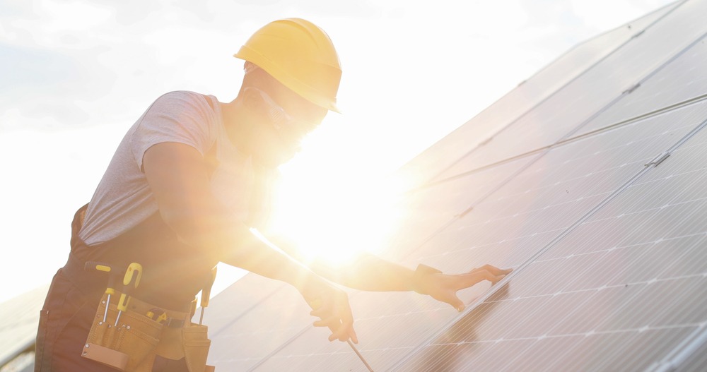 A worker wearing a hard hat and safety goggles is installing or maintaining solar panels under bright sunlight. The worker is using tools from a tool belt and is positioned in front of an array of solar panels.