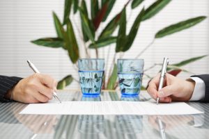 man and woman signing divorce document on a glass table with two glasses of water and a plant in the background.