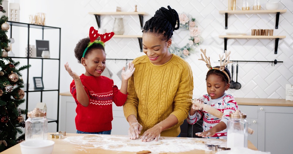 Cheerful African American woman in apron with little kids standing at table in home Christmassy decorated kitchen and having fun while making xmas cookies.