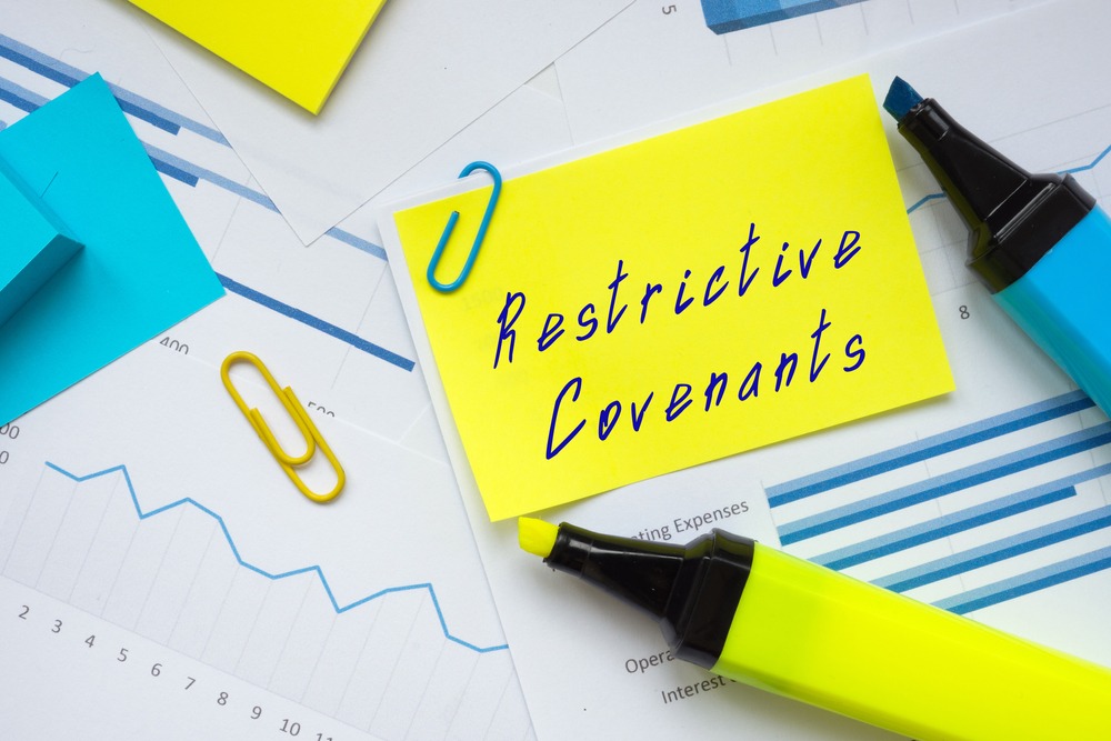 The words restrictive covenants written on a yellow sticky note surrounded by highlighters, paper clips and papers with charts and graphs