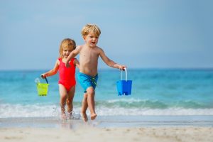 Little boy and a girl, siblings run barefoot on sea beach from waves carry toy buckets with water