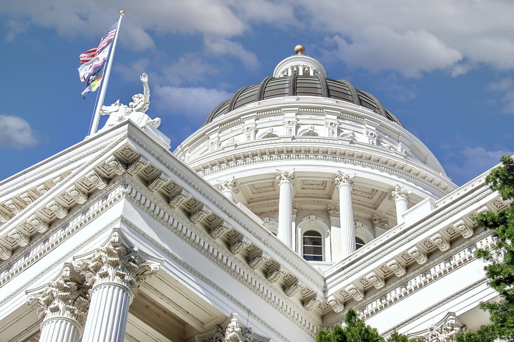 Close-up view of the California State Capitol building's dome, featuring intricate architectural details and columns. The American flag and California state flag are visible, flying against a partly cloudy sky.