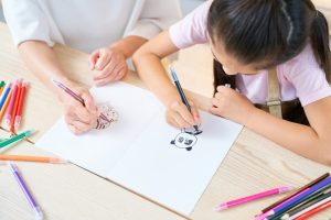 Parent and child drawing on a white piece of paper with colored pencils scattered on a wooden table