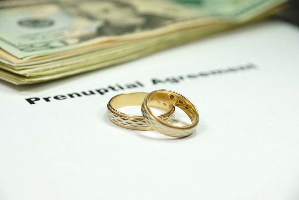 Two golden rings and a stack of US 20 dollar bills sitting on top of a white piece of paper with Prenuptial Agreement in black type.